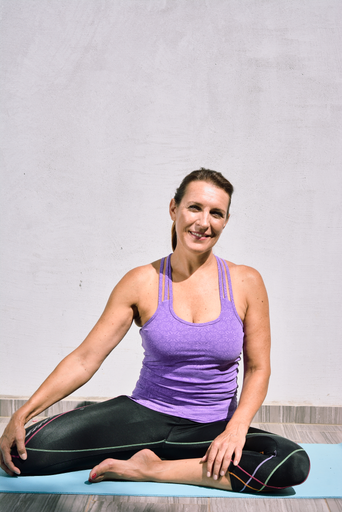 Ženska sedi na podlogi za pilates, gleda v stran in razteguje noge.